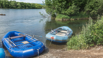 Inflatable boats for rafting stand by the river bank. Ripples on clear blue water. Green vegetation around. Kamchatka