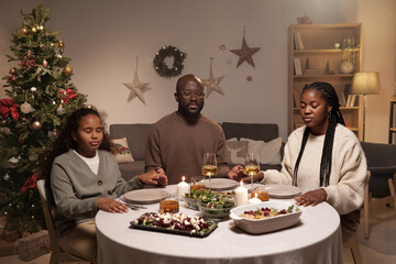 Fototapeta na wymiar Contemporary Christian family of three praying before festive dinner while sitting by table served with homemade food