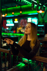 Young attractive woman with long blond hair in a nightclub restaurant-bar in neon light green color stands. Fashion model in neon light green color.