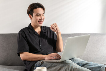 Joyful young Asian young man sitting on couch smile is celebrating win victory and showing yes gesture during online education at home, getting a new job offer or make good selling win happy concept