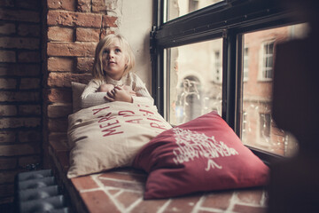 A little girl sits on a brick windowsill surrounded by pillows with Christmas inscriptions and makes wishes to Santa