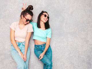 Two young beautiful smiling blond hipster female in trendy summer clothes. Sexy carefree women posing near grey wall in studio. Positive models having fun. Cheerful and happy. In sunglasses