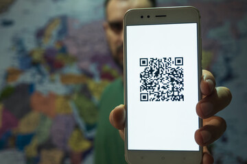Close-up of a smartphone screen with a QR code of the covid-19 digital vaccination certificate. In the background is a man against the background of a world map