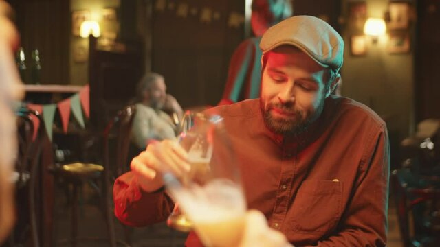 Over-the-shoulder shot of two Caucasian men wearing casual clothes having fun in pub clinking glasses with beer, drinking it and discussing news