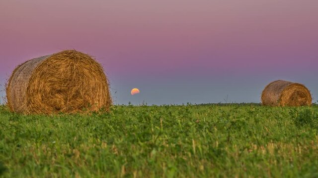 5k video of 2 hay bails of straw in a green feild and movement of the moon in the background. Timelapse from evening to night.