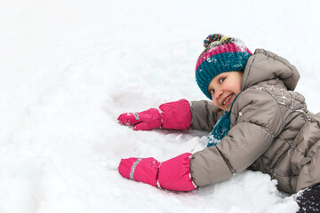 Fototapeta na wymiar Winter walking activity. The girl is lying in the snow and playing. Child kid in a winter suit, knitted hat and scarf and waterproof gloves. looking in camera