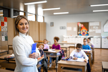 Portrait of school teacher with notebook standing in classroom and children sitting in background.