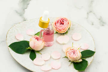 a bottle of rose oil filled with a pipette of aromatic rose oil on a ceramic plate and a white marble background. close angle.