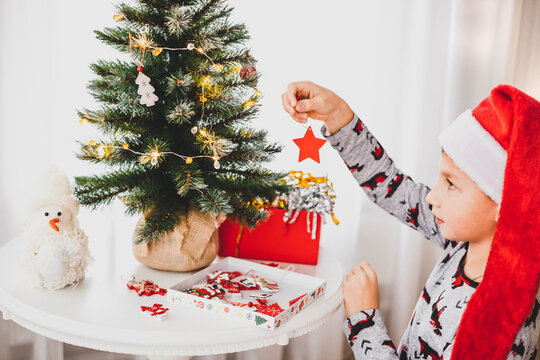 Christmas And New Year Vacation. Child Boy Decorating Little Christmas Tree During Winter Holidays. Teenager In Pajamas And Red Santa Hat Hanging Decorative Wooden Toy On Fir Branches.