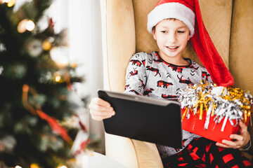 Happy teenager boy in pajamas and red santa hat with tablet having video chat at home. Smiling...