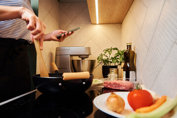 Woman use smartphone while cooking bolognese sauce in pan on induction stove in the kitchen
