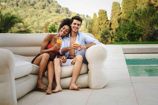 happy couple on vacation drinking cocktails sitting by the swimming pool - multicultural couple of young people drinking fruit juice poolside. - Powered by Adobe