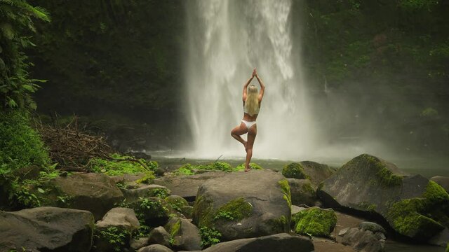 Blond woman in tree yoga pose balance with powerful Nungnung waterfall