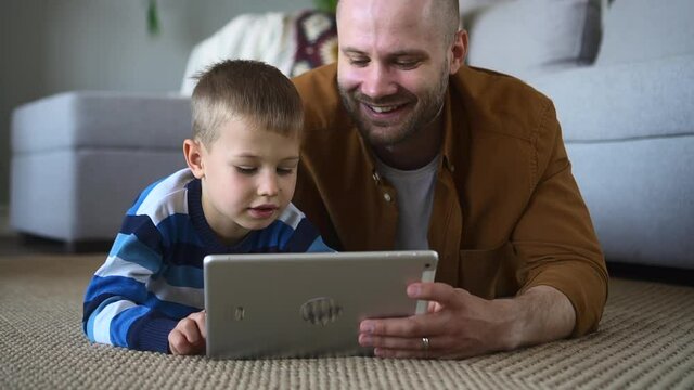 Father and son play online games. Happy little child does interesting tasks on contemporary tablet spbd lying with careful bearded man on floor in living room