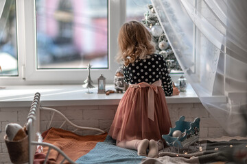 A little girl is sitting on the bed by the window and decorating a small tree with tiny Christmas toys. Happy healthy child celebrating a traditional family holiday. Adorable baby.