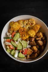 Vegan lunch bowl: baked cauliflower, tofu, tomato and cucumber salad. Dark wooden background. 