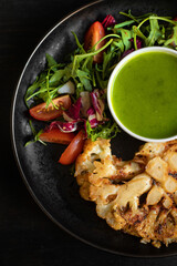 Cauliflower steak with vegetable salad and pesto sauce on a black plate, black background. Close top view.
