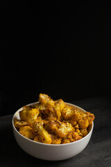 Baked cauliflower in white bowl on dark wooden desk, black background.