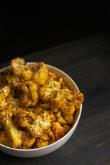 Baked cauliflower in white bowl on dark wooden desk, black background. Top view.