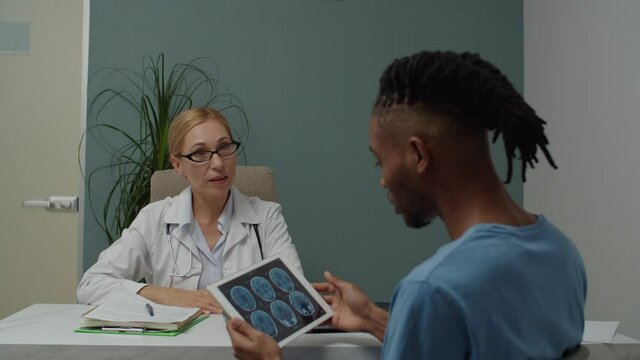 Young adult male patient in casual clothing using tablet, looking at MRI digital image, questioning female doctor about result, diagnosis indoors while having physician's appointment in clinic
