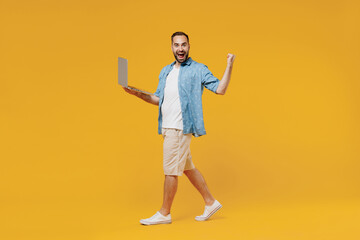 Full body young smiling happy fun caucasian man 20s wearing blue shirt white t-shirt hold use work on laptop pc computer do winner gesture walk go isolated on plain yellow background studio portrait