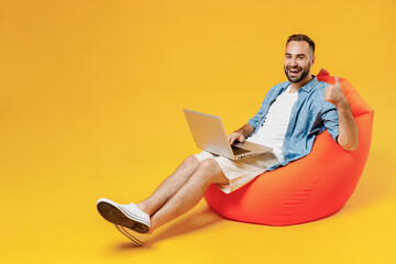 Full body young smiling fun cool happy man wear blue shirt white t-shirt sit in bag chair hold use work on laptop pc computer show thumb up gesture isolated on plain yellow background studio portrait