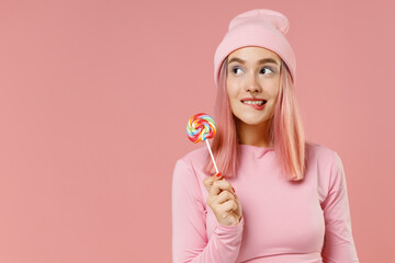 Young minded woman 20s with bright dyed rose hair in rosy top shirt hat hold sweet candy lick eat lollipop look aside isolated on plain light pastel pink background. People lifestyle fashion concept.