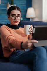 Student with glasses on couch holding cup looking at tablet display. Influencer on sofa in videocall conference while drinking coffee or tea. Content creator streaming video on digital device.