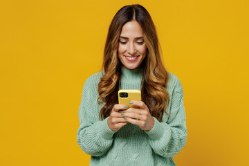 Young smiling woman 30s wearing green knitted sweater hold in hand use mobile cell phone chatting browsing internet isolated on plain yellow color background studio portrait. People lifestyle concept.
