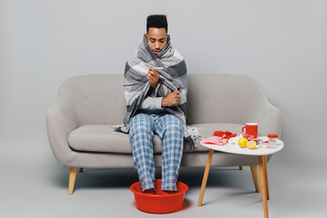 Young ailing african american man in grey plaid scarf sit on sofa soaking feet in warm water isolated on plain gray background studio portrait. Healthy lifestyle ill sick disease treatment concept