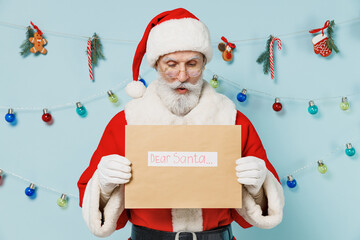 Old amazed Santa Claus man 50s wears Christmas hat red suit hold reading letter with dear santa title text isolated on plain blue background studio. Happy New Year 2022 merry ho x-mas holiday concept.