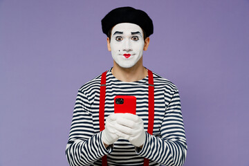Charismatic amazing fun young mime man with white face mask wears striped shirt beret look camera hold in hand use mobile cell phone isolated on plain pastel light violet background studio portrait.