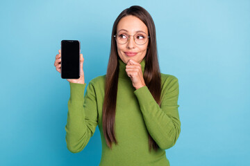 Photo of cute thoughtful young woman wear green turtleneck glasses holding modern device empty space arm chin isolated blue color background