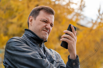 Sad millennial guy looks at phone screen in surprise and stands outside at autumn. Disgusted and...