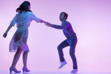Studio shot of young man and woman in vintage retro style outfits dancing lindy hop isolated on lilac color background in neon light