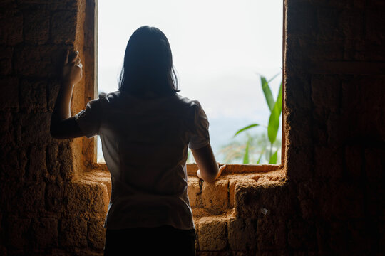 Silhouette poor young woman looking through the window at old home.