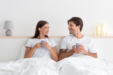Cheerful happy young european husband and wife look at each other, hold cups in bed at bedroom interior