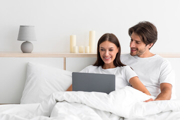 Smiling attractive young caucasian husband with stubble hugs wife sitting on bed, use laptop to surf in internet