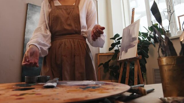 Art studio - young woman artist puts undone painting on the easel and continue the work