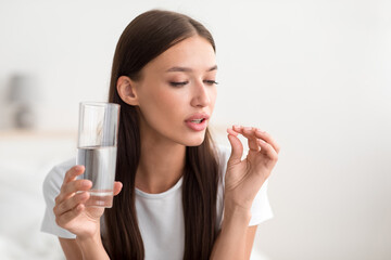 Millennial pretty caucasian female hold pill and glass of water in bedroom interior