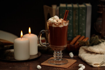 Cocoa or hot chocolate with marshmallows and a cinnamon stick in a tall glass glass. Against the background of books, burning candles and a knitted plaid. Close-up