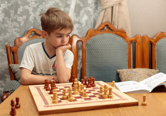 A blond-haired boy in front of a chess board, pondering a problem from a textbook. Portrait,...
