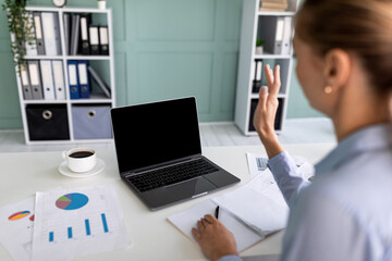 Remote meeting. Over shoulder view of businesswoman sitting at desk and talking via laptop, waving at webcam