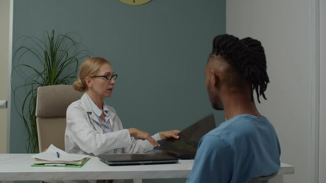 Mature female doctor holding MRI image, interpreting results on picture, explaining diagnosis to young male patient indoors. Healthcare specialist holding medical appointment, talking with case