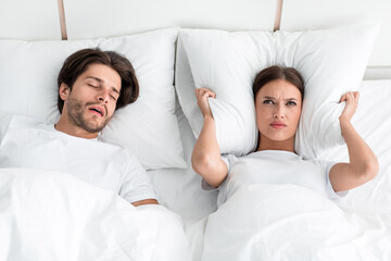 Attractive millennial european guy with stubble sleeps on bed, angry upset wife covers ears with...