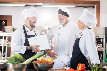 Asian chef holding cookbook and talking to smiling colleagues near food in kitchen