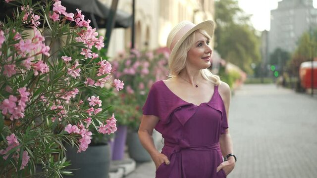 Elegant confident lady walking out to city street passing flower pots and looking at camera smiling. Portrait of happy slim beautiful Caucasian woman strolling in the morning in European town posing