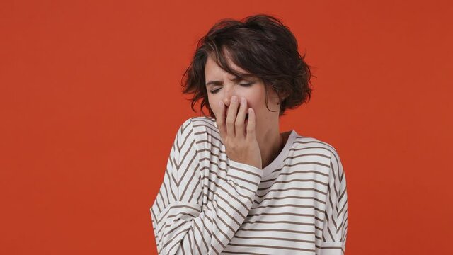 Tired young brunette woman 20s years old wear striped shirt did not get enough sleep last night after party and barely got up in the morning yawning isolated on plain orange background studio portrait
