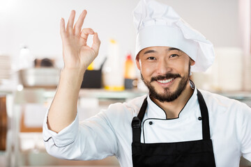 Portrait of smiling asian chef in uniform showing okay gesture in kitchen