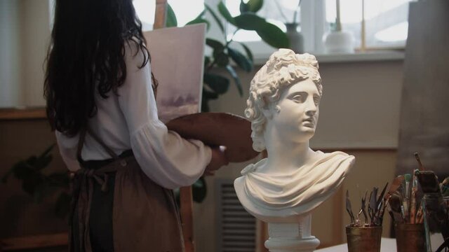 Young woman artist with long hair drawing a painting - marble bust on the foreground